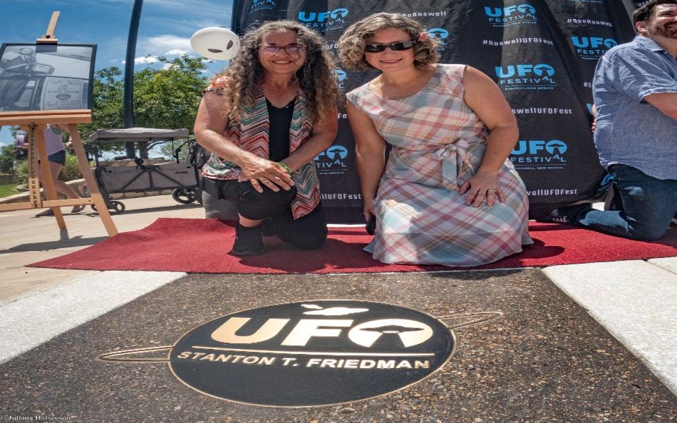 Two women crouched for a picture with a floor title dedicated to the Roswell, New Mexico UFO Festival.